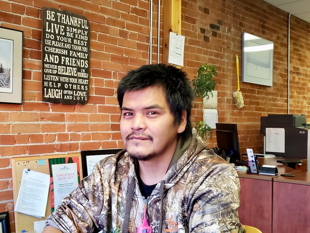 A man with black hair and wearing a light-coloured patterned hoodie sits at a table in a coffee shop, looking at the camera. There&rsquo;s a red brick wall behind him.