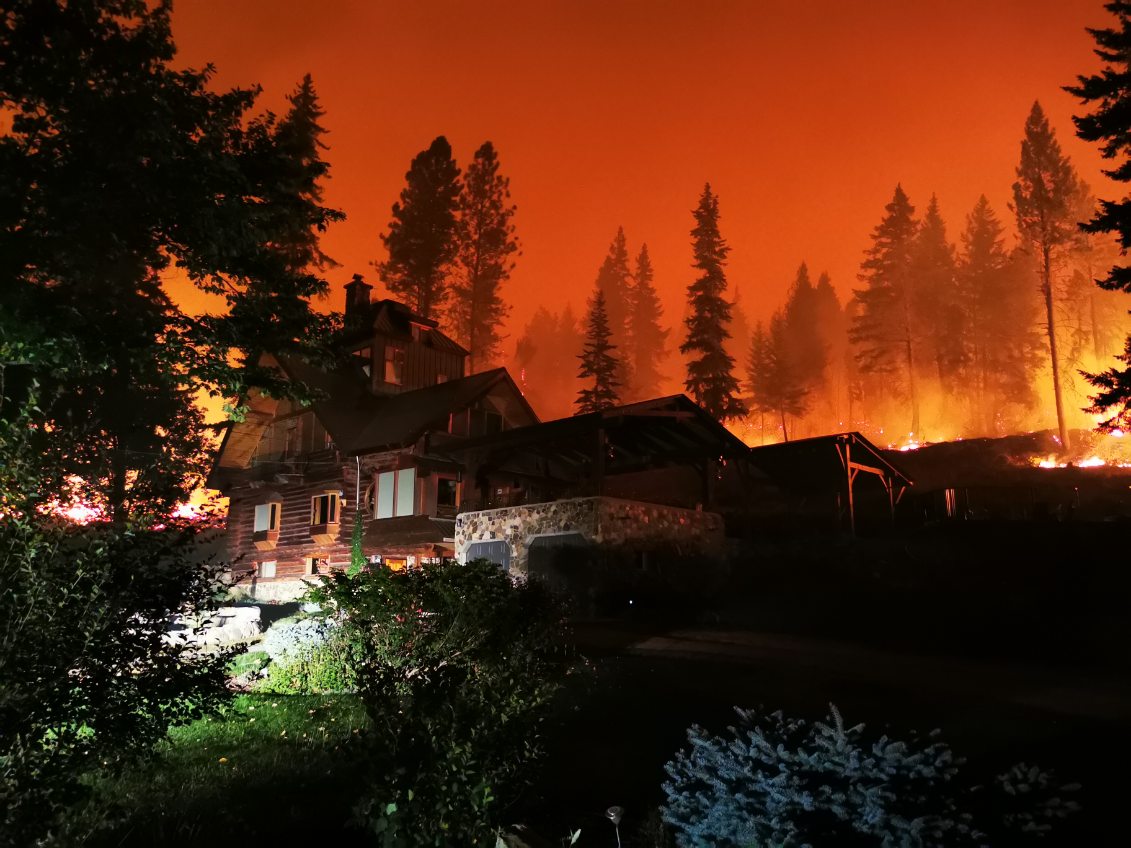 It’s nighttime and a home sitting on a terraced slope is illuminated at the front with wildfire filling the sky behind.