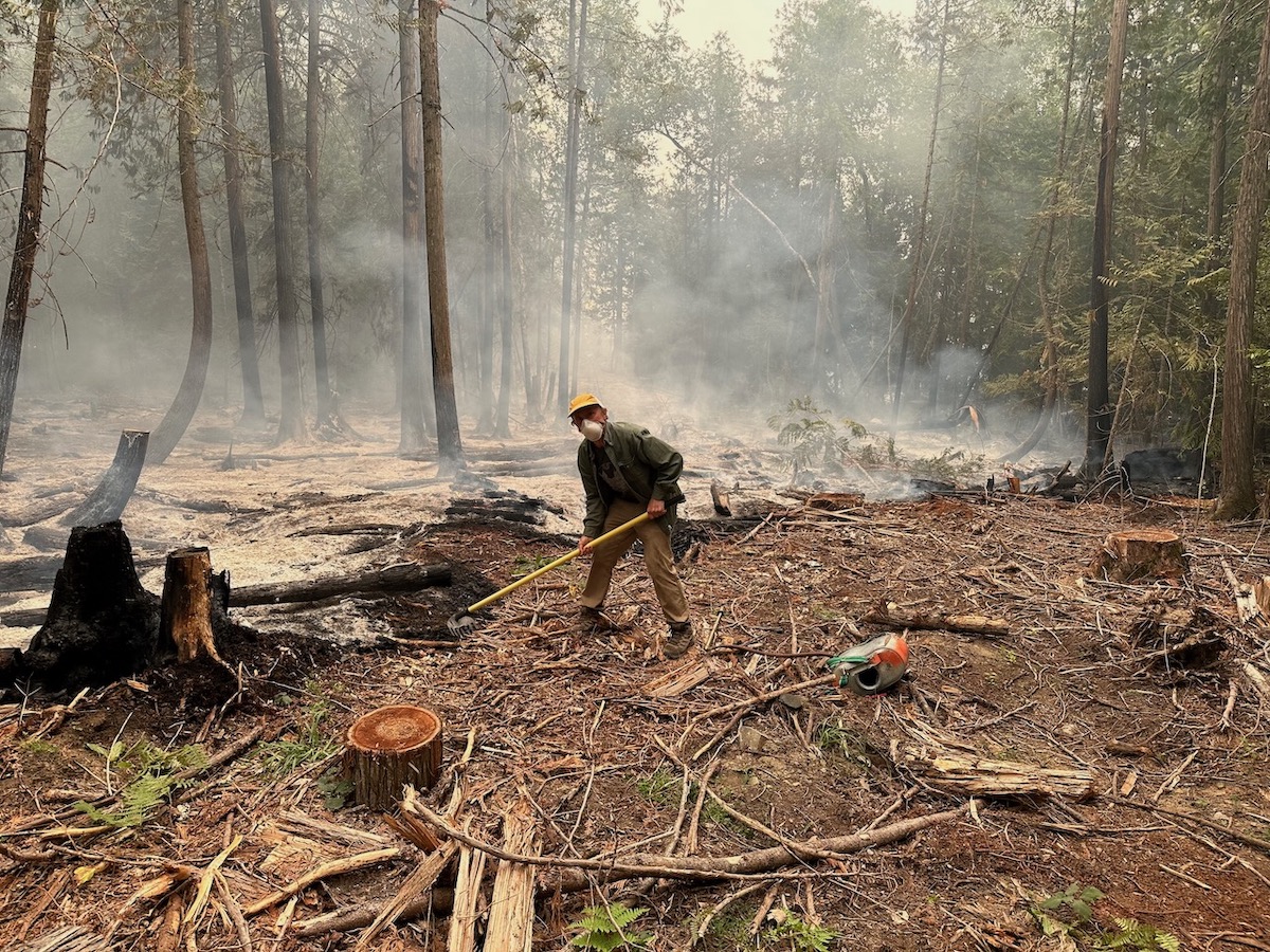 A man wearing a face mask rakes the ground. Behind him, the ground is smouldering and covered in ash.