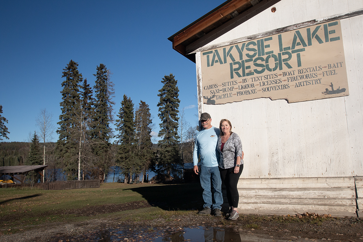 A man and a woman stand with their arms around each other under a sign, which has a corner missing, that says Takysie Lake Resort. A lake can be seen in the background.