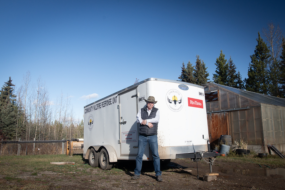 A man with his arms crossed in front of him stands in front of a white trailer with blue sky behind.