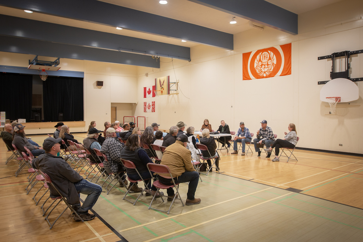 In a large gymnasium, people sit in several rows of chairs looking at five people who sit facing them, speaking to the audience.