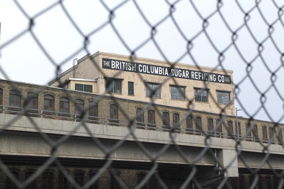 A photo taken through a chain-link fence shows a long one-storey brick industrial building. Behind it rises a taller building with a painted section on top that says "The British Columbia Sugar Refinery Company Ltd."