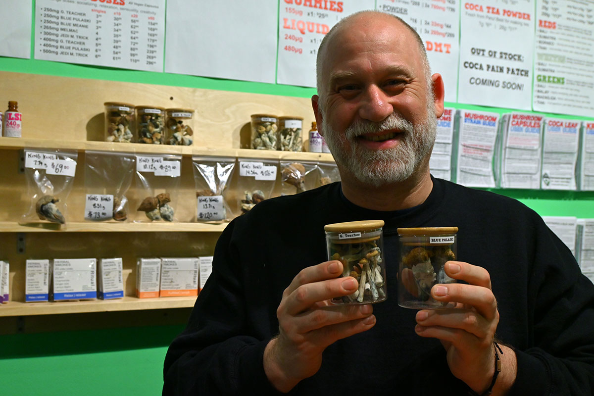 A light-skinned man with grey hair and a beard holds two jars of psychedelic mushrooms.