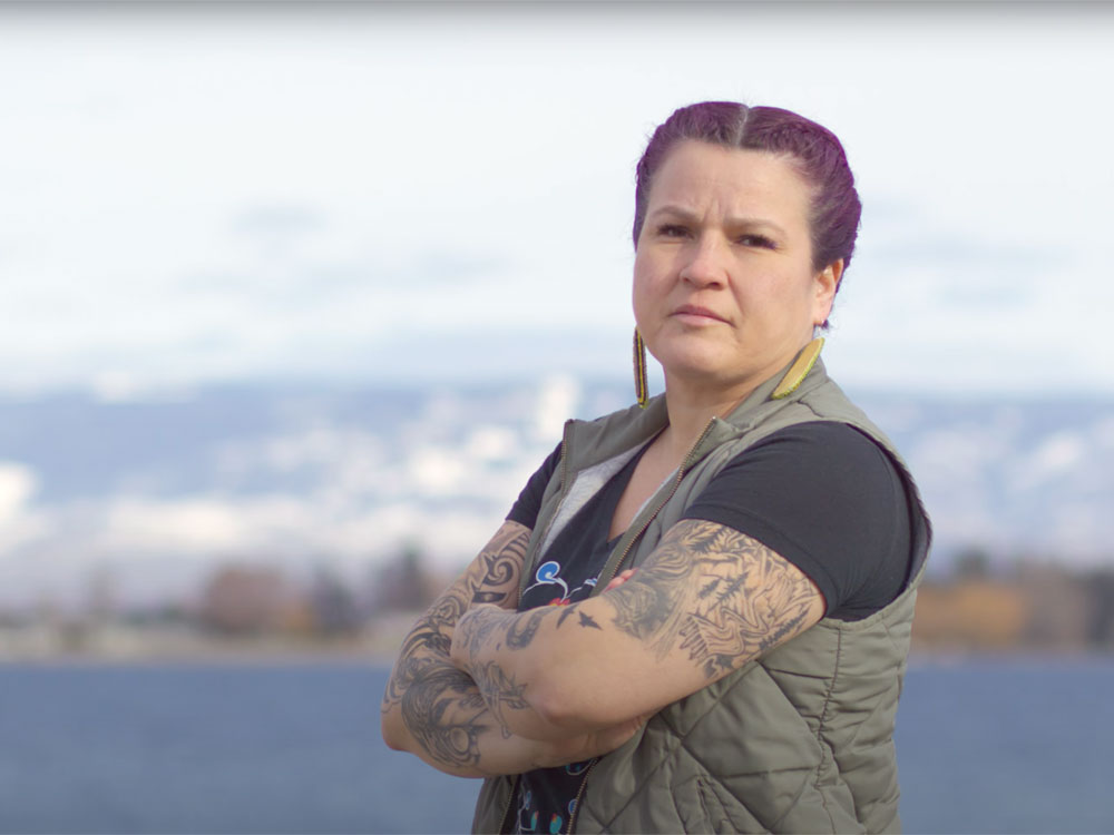Heather Spence, a lighter-skinned Indigenous person with purple hair in braids, stands in front of the ocean with arms crossed.