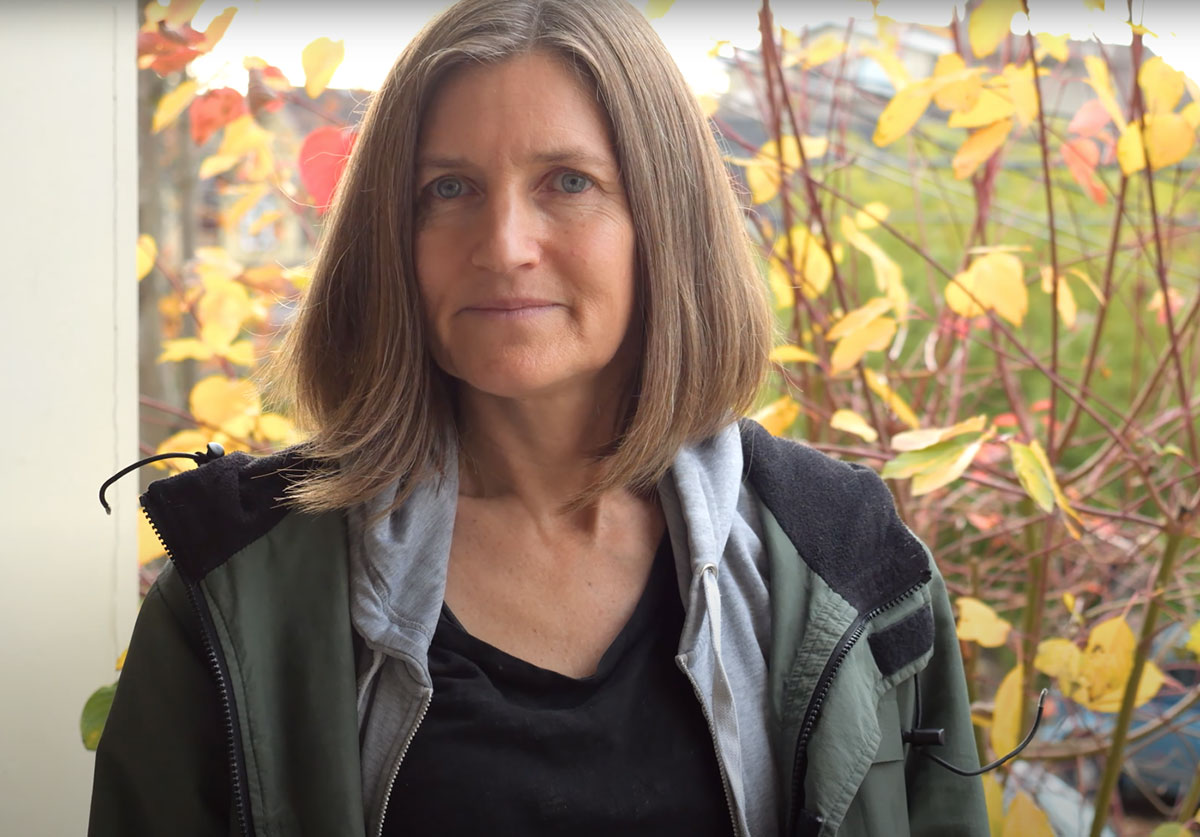 Keri Guelke, a white woman with her light brown hair cut in a shoulder-length bob, looks directly at the camera. She is wearing a coat and sweatshirt and sitting in front of a window.