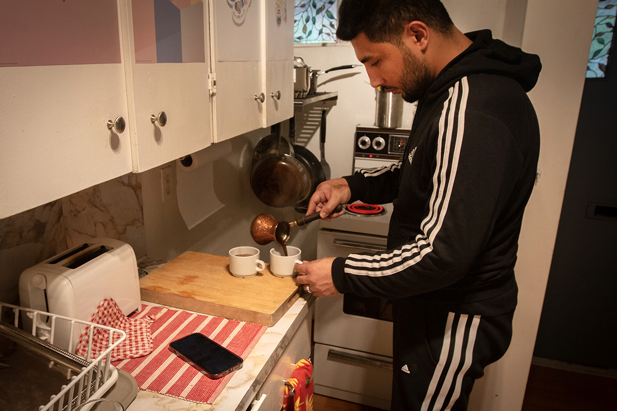 A man with dark skin tone, hair and beard uses a small copper pot to pour coffee into two white mugs that sit on a narrow countertop. There are white kitchen cabinets above the countertop and a stove sits behind him.