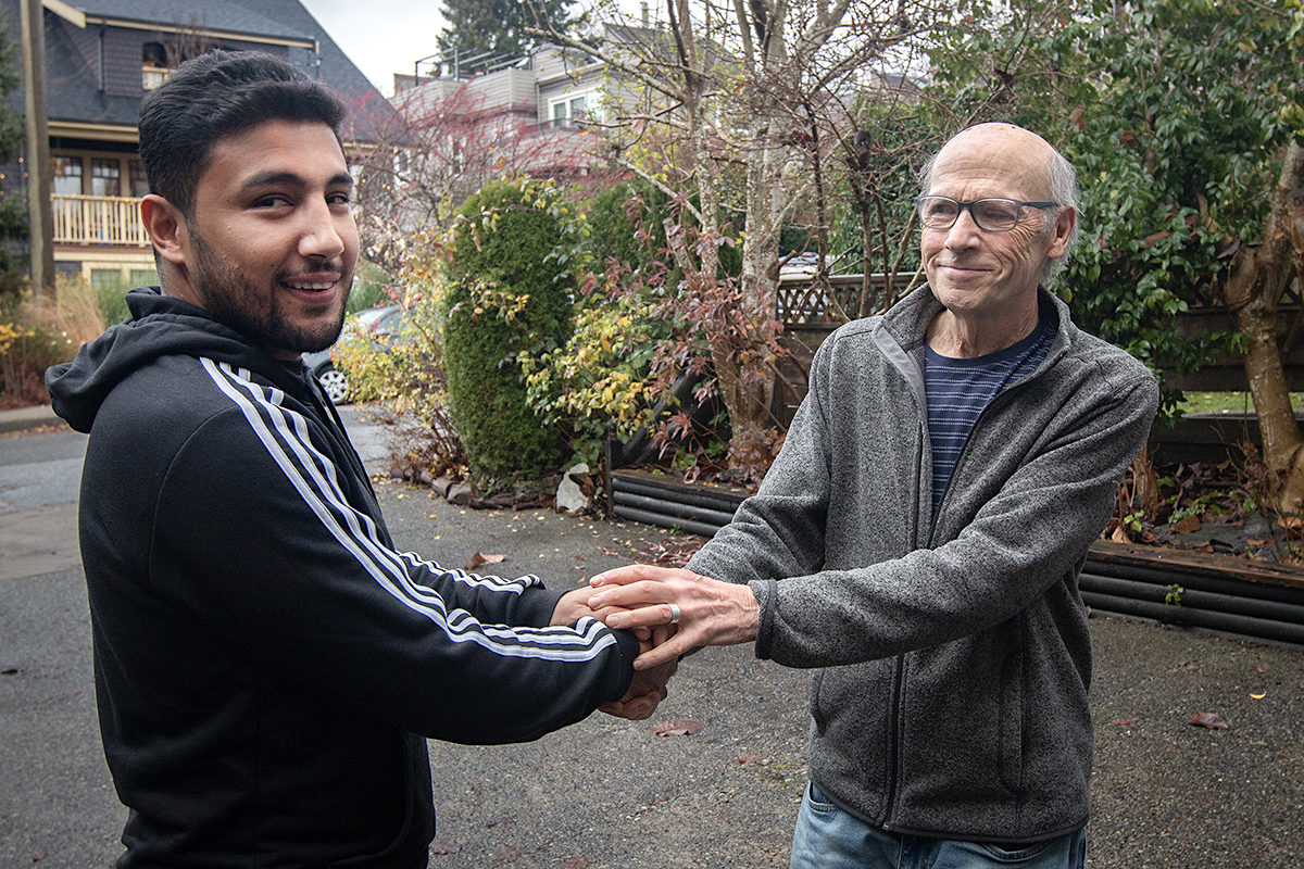Two men stand outside with shrubbery behind them. They face each other with their hands clasped. The man on the left looks at the camera and smiles. He has dark skin tone and dark hair. The man on the right is older, balding and wears glasses. He looks at the other man and is smiling.