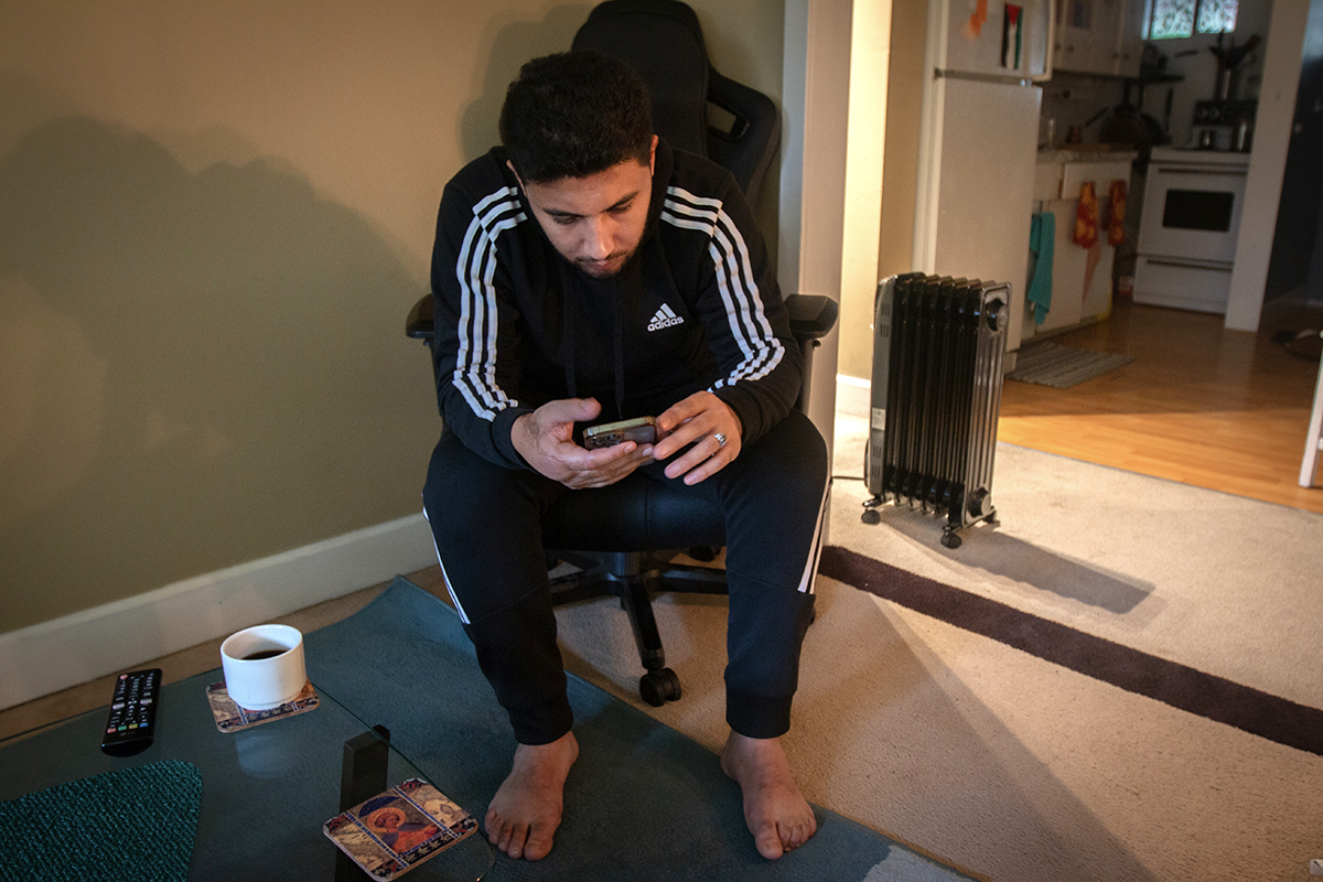 A man with dark skin tone, hair and beard sits in a black chair and looks down at a cellphone in his hands.
