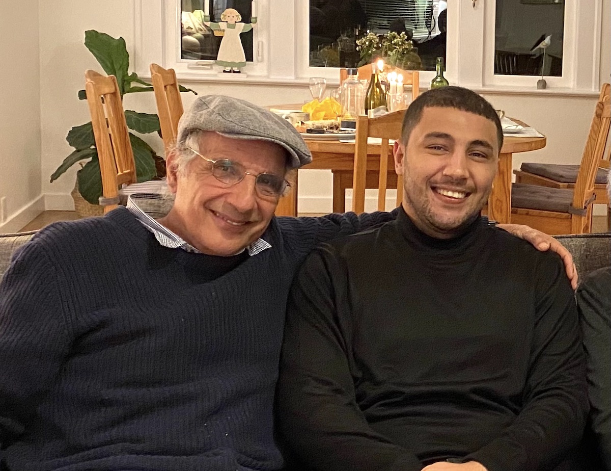 Two men sit side by side. The man on the left is middle-aged and wears a hat and glasses. He has an arm around the young man on the right, who has dark skin tone and very short dark hair. Both men are smiling.