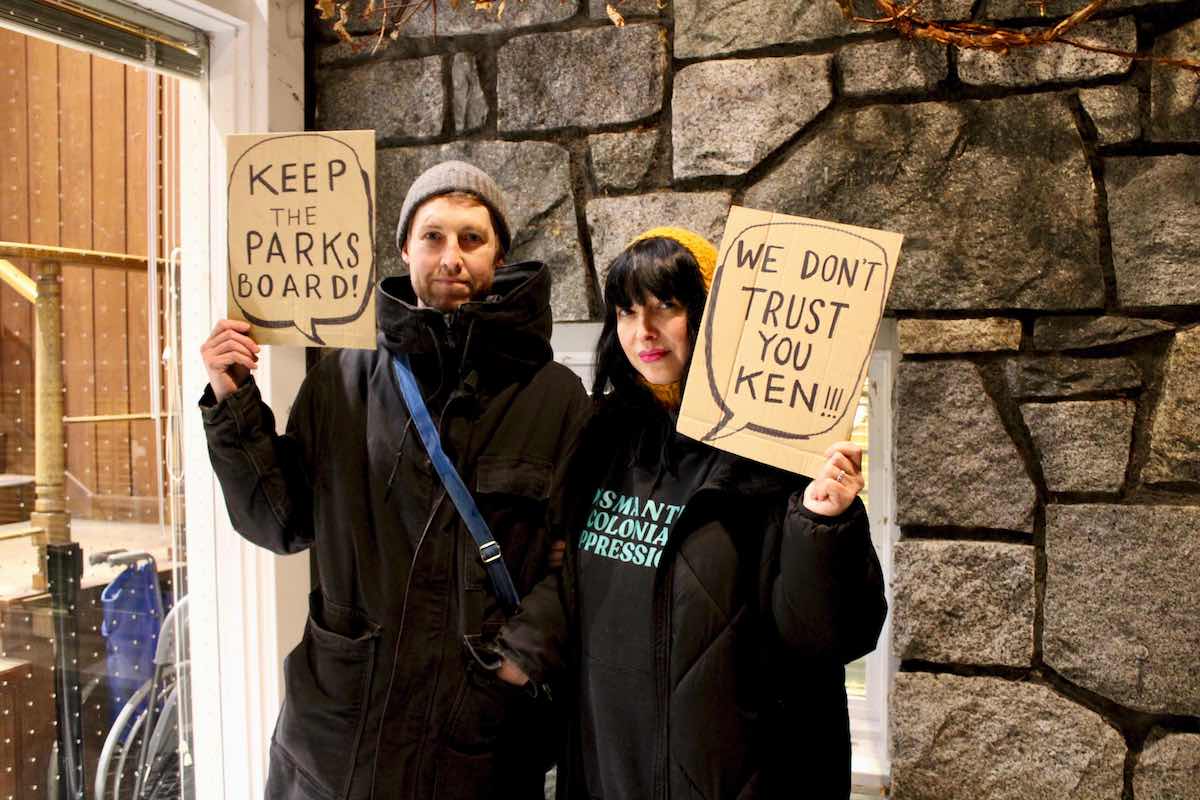 Two young people dressed in black hold cardboard signs that say 'Keep the parks board' and 'We don&rsquo;t trust you Ken.'
