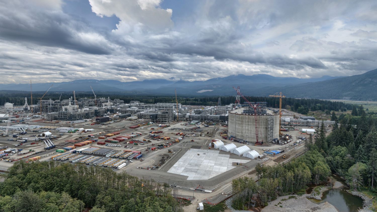 The photo shows a sprawling industrial complex under construction, with dozens of large buildings, cranes and a giant round concrete structure. It&rsquo;s a cloudy day, and mountains are visible in the background.