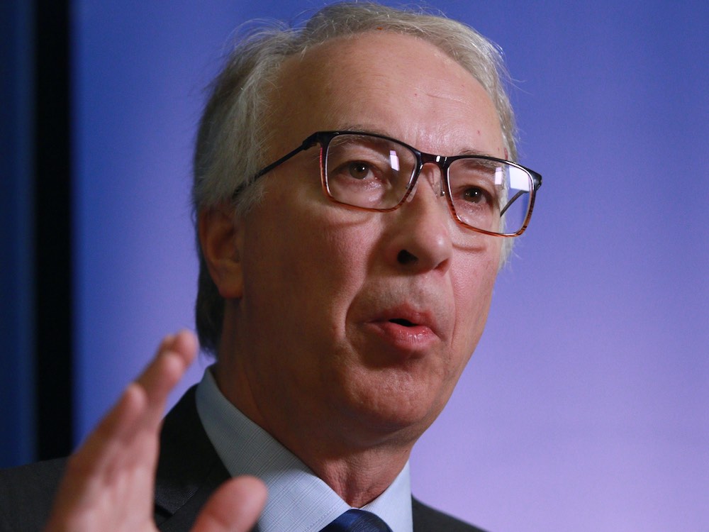 A 60-ish white man with grey hair and dark-framed glasses gestures as he speaks to an unseen audience. He wears a blue shirt and dark blue tie.