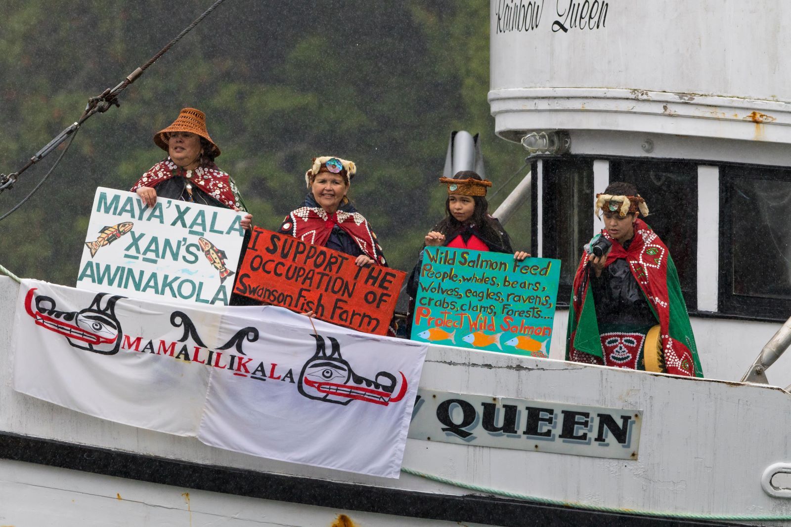 A group of Indigenous people, two older and two younger, dress in traditional garb and hold signs that carry messages including 'I support the occupation of Swanson Fish Farm.' One of them is looking into a small camera.