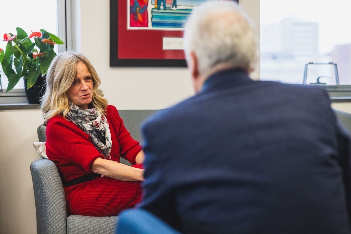 Rachel Notley, a woman with a light skin tone and medium-length blond hair, wearing a red dress and a white and black patterned scarf, sits on a grey couch in mid-speech, opposite a man with grey hair and a dark blue jacket with his back to the camera.