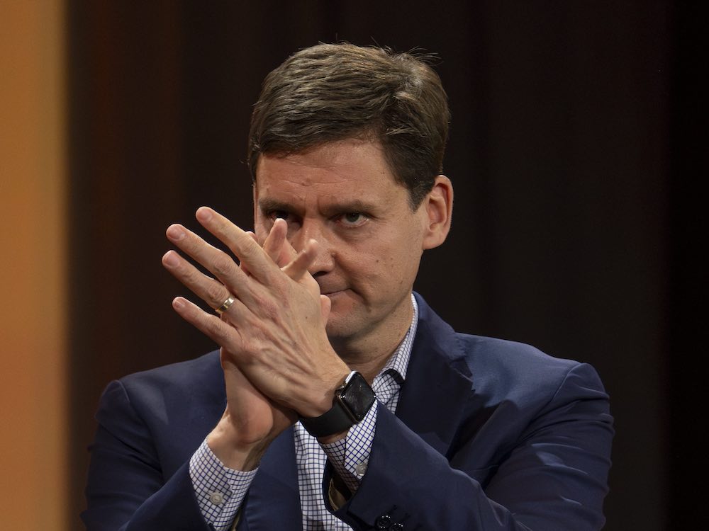 A 47-year-old white man with medium-length dark hair, wearing a blue suit and a white and blue checked short, looks to the side and appears to be applauding.