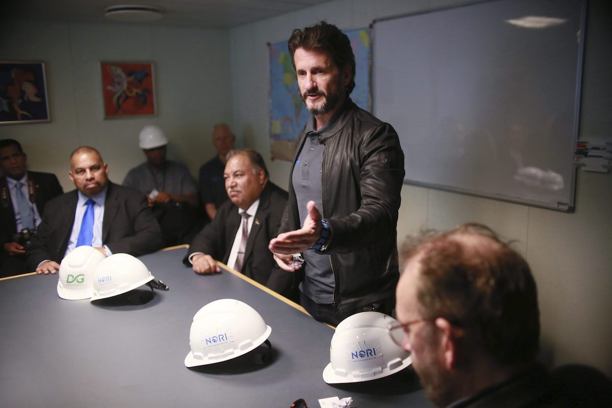 A middle-aged white man with stylish brown hair and a greying beard speaks at a table with other men in suits sitting around. He is slim, wearing a grey polo shirt and brown leather jacket.