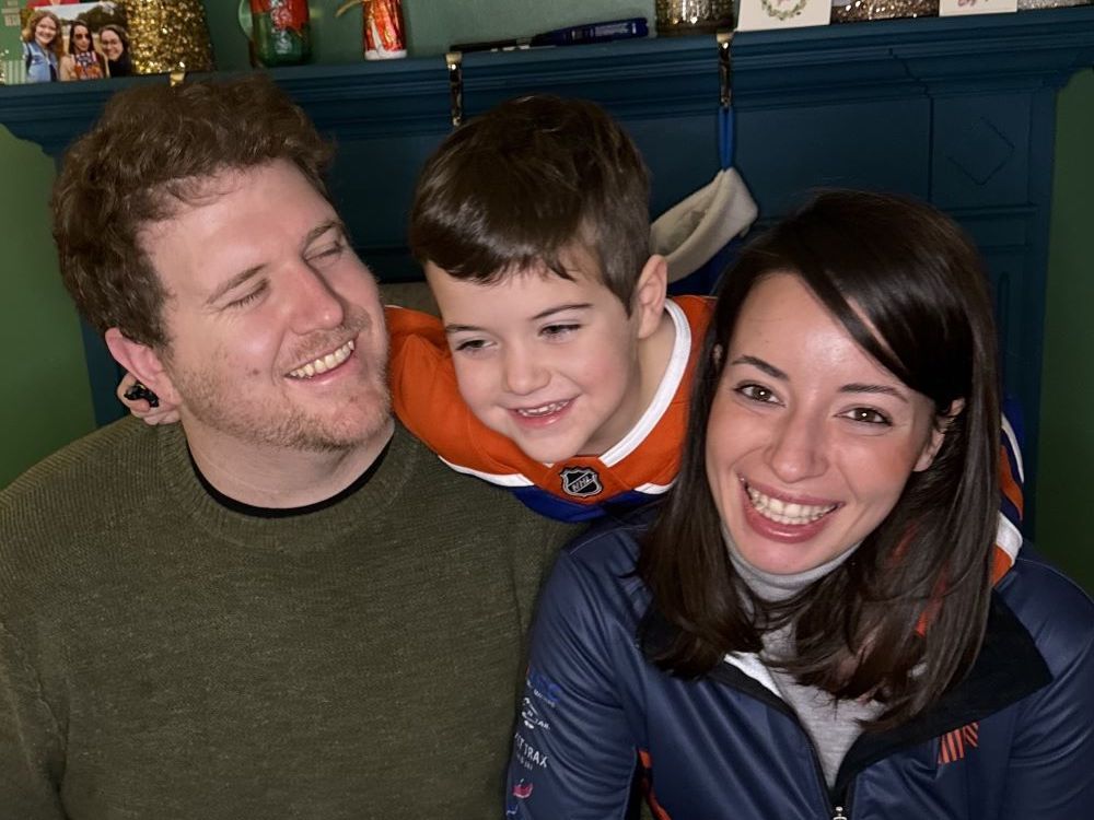 A family of three poses in front of a mantel. At left, a man with light skin tone and brown hair smiles at his five-year-old son, a boy with dark brown hair and light skin tone, at centre, next to a woman with dark hair and eyes and medium-light skin tone, at right.