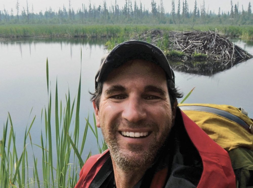 A bearded light-skinned man wearing a ball cap smiles at the camera. Behind him sits a structure made of stacked wood &mdash; a beaver lodge.