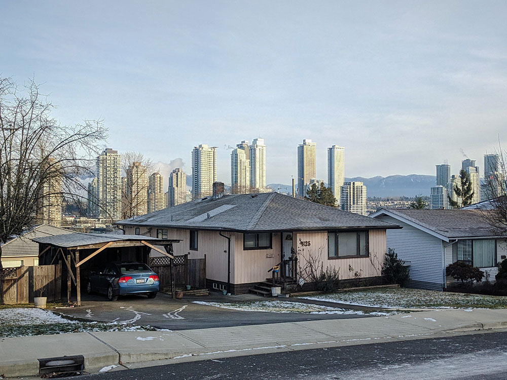 It’s a snowy day and there is a skyline of new glass towers looming over a tired-looking house built in the 1950s.