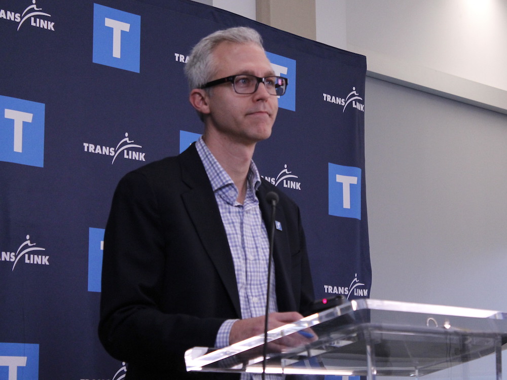An early middle-aged white man with short grey hair, stylish glasses and a dark jacket and checked shirt stands at a podium.