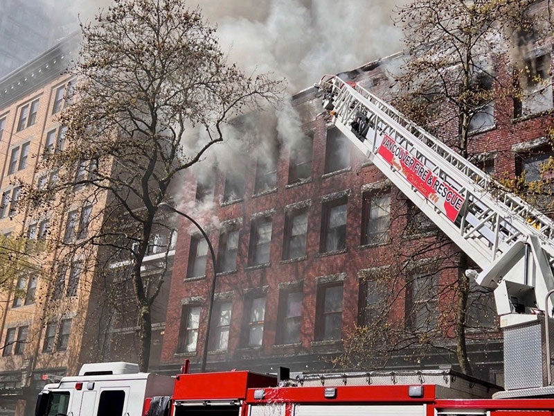 The top section of a red fire truck and ladder as smoke pours from the upper windows of a four-storey red brick building.