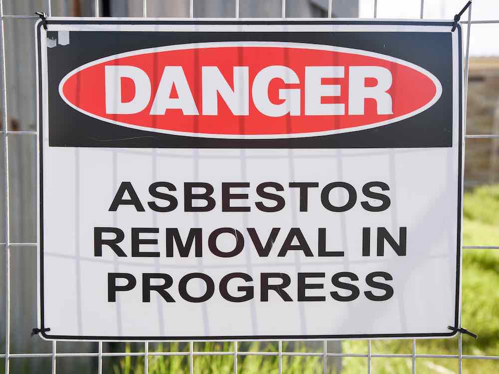 A close-up of a sign fastened to a wire fence, with a building in the background. White letters in a red oval say 'DANGER.' Beneath that, black letters on a white background say 'ASBESTOS REMOVAL IN PROGRESS.'