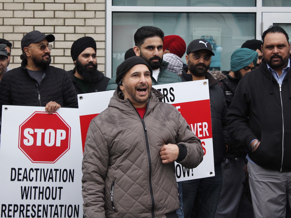 An Indo-Canadian man in a brown jacket and black toque is smiling and speaking in front of a group of other Indo-Canadian men. A sign to his right says 'STOP DEACTIVATION WITHOUT REPRESENTATION.'