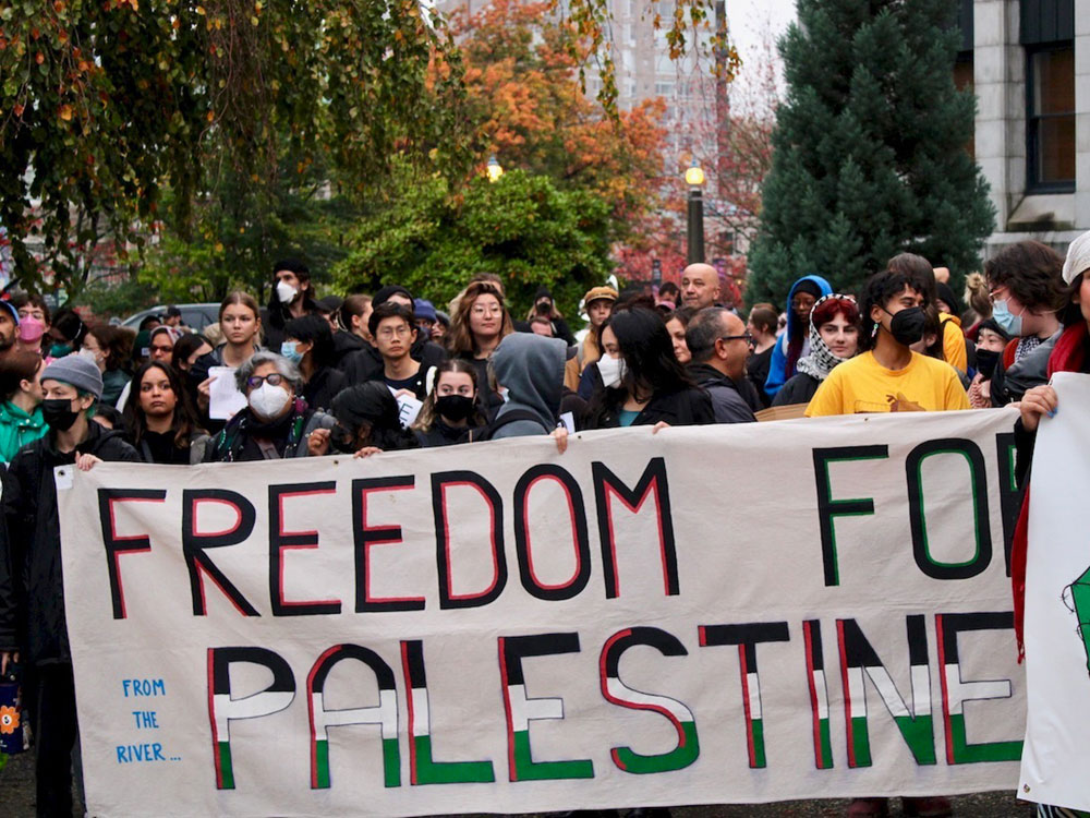 A group of people hold a banner that says 'Freedom for Palestine.' A larger crowd is gathered beside and behind them.