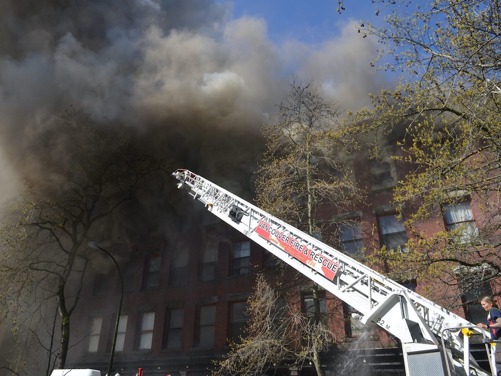 Clouds of smoke in shades of grey billow from the top floor of a red brick building. The silver ladder from a fire truck stretches toward the smoke from the lower right, under a blue sky.