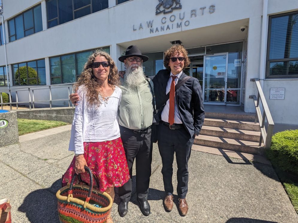 Three people stand in front of the Nanaimo Law Courts building. From left, a woman with light skin and long curly brown hair wearing a white peasant blouse and red patterned skirt; a man with light skin and a long grey beard wearing a black fedora, a moss green button-up shirt and black pants; and a man with light skin and curly brown hair wearing a dark casual suit and red tie.
