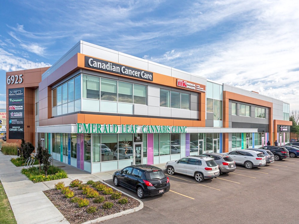 A two-storey office building has a 'Canadian Cancer Care' sign on the top floor, above the 'Emerald Leaf Cannabis Clinic.' All but one of the parking spaces are full in the lot in front of the building.