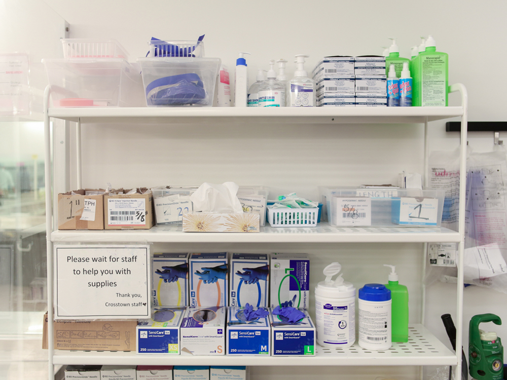 Harm reduction supplies on a shelving unit. They are well-organized and labelled. A sign says &lsquo;Please wait for staff to help you with supplies.&rsquo;