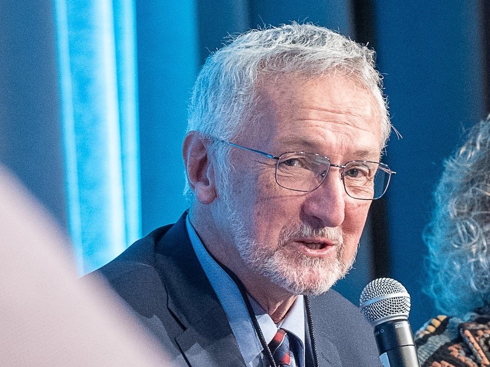 A white-haired white man of late middle age with a neatly trimmed beard and wire-rimmed glasses leans forward while speaking into a microphone.