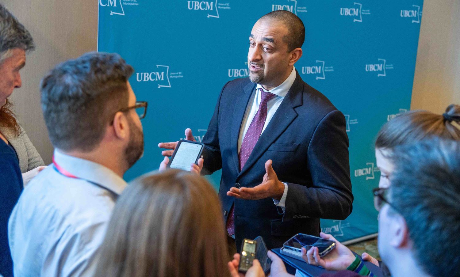 Ravi Kahlon is a medium-skinned man wearing a navy suit over a white shirt and burgundy tie. He stands against a wall with a wide blue Union of BC Municipalities banner before a group of journalists holding smartphones and voice recorders towards him. Kahlon is mid-speech with his hands raised.