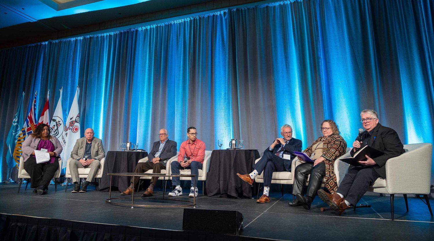 Seven people sit on off-white low-slung upholstered chairs across a wide stage in front of a row of blue curtains. Small tables with black tablecloths and pitchers of water are set between some of the chairs. A person on the far right in black pants, a black blazer and a dark button-down shirt with short grey hair and glasses holds a microphone, addressing the crowd.