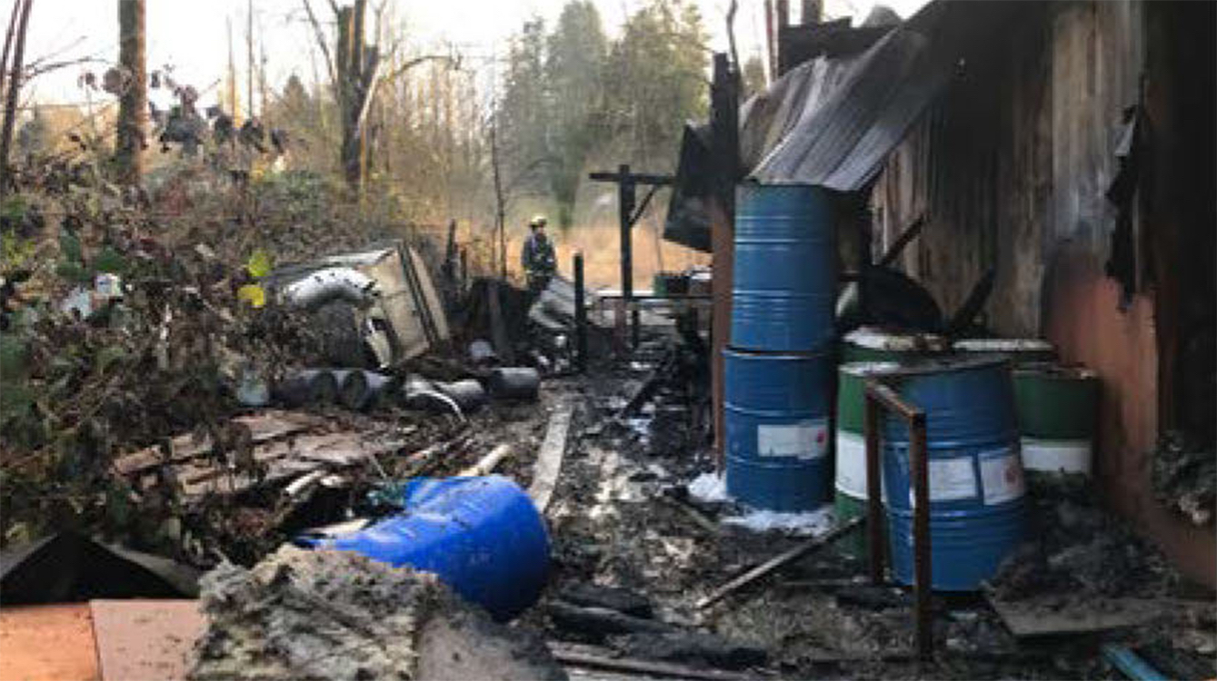 A photo showing metal barrels and other debris strewn about, with a person in firefighting gear standing at the centre.