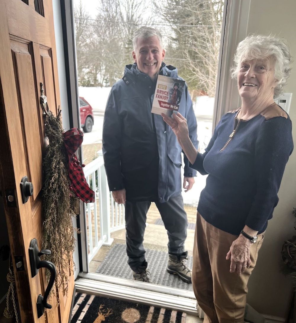 An elderly light-skinned woman stands at her door holding a flyer that reads 'Vote Bruce Fanjoy.' Fanjoy, a light-skinned man with grey hair, stands outside the door.