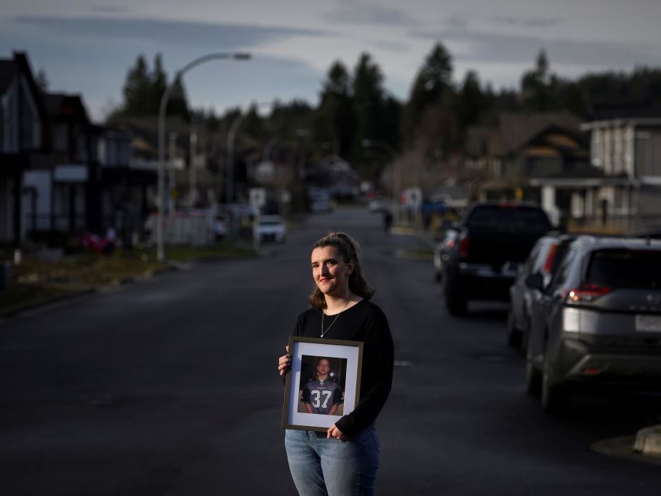 A light-skinned woman stands in a street, illuminated by sunlight against an overcast sky. She is holding a photo of a man in a football shirt.
