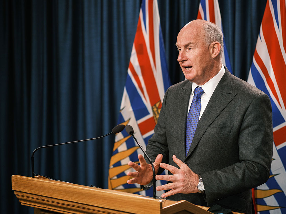 A 64-year-old light-skinned man stands at a podium wearing a dark suit, white shirt and blue tie.
