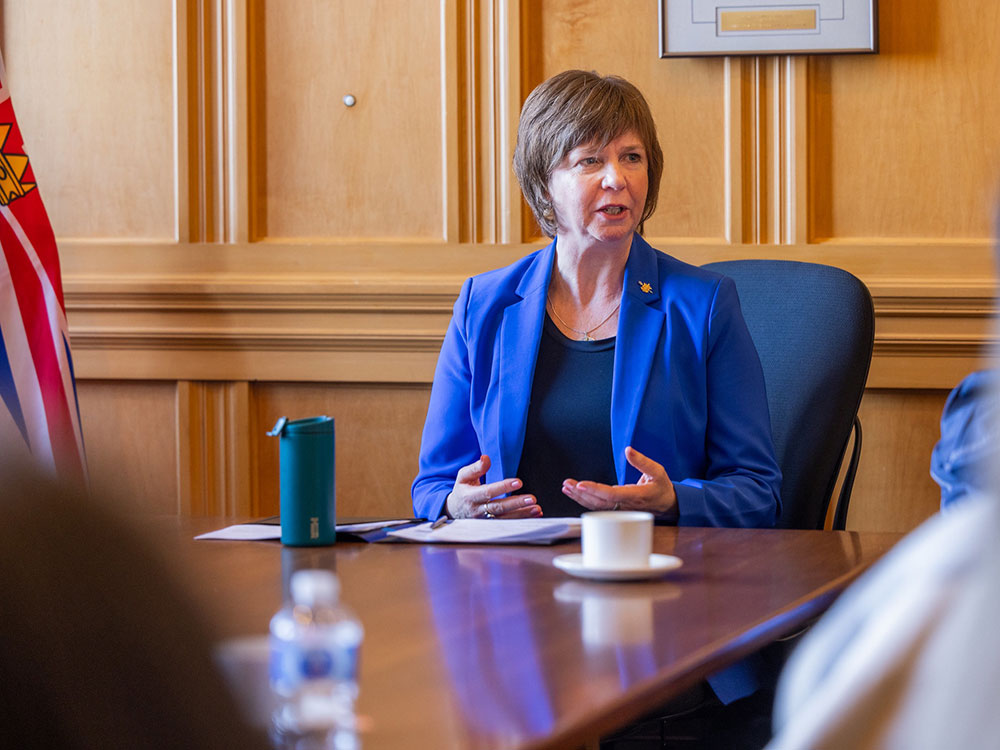 A 57-year-old light-skinned woman sits at a conference table. She has short brown hair and wears a blue jacket and shirt.