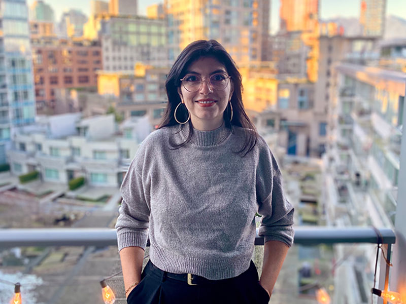 A young light-skinned woman with long dark hair leans against a balcony railing and smiles at the camera. She wears a grey sweater and gold hoop earrings.