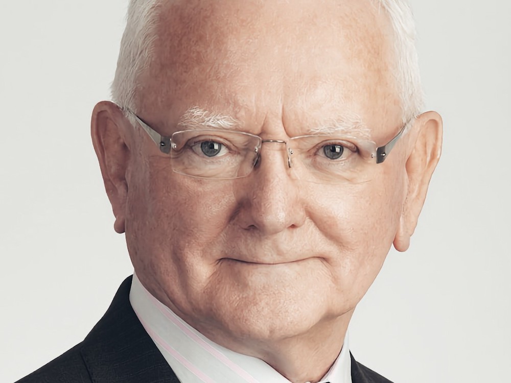 A man with a light skin tone and white hair is wearing glasses and a suit, and looking directly into the camera lens.