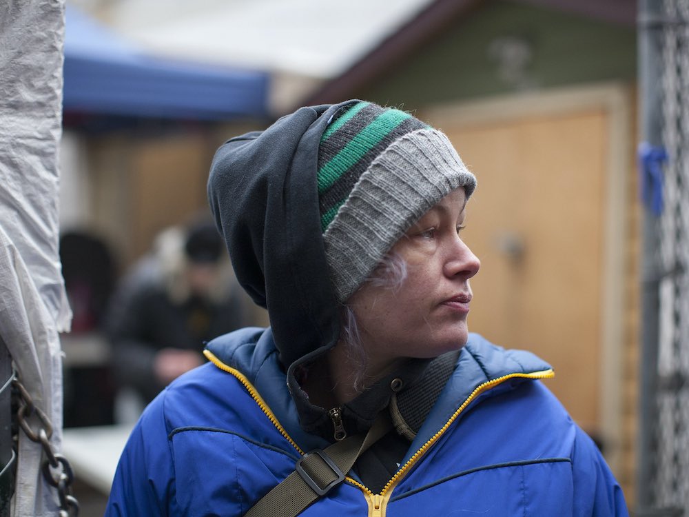 A woman looks purposely to her to left. She is beside a chain-link fence, wearing a toque, black hoodie and bright blue jacket.