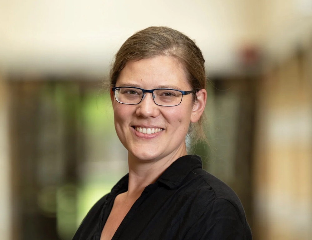 A light-skinned woman with tied-back dark blond hair and glasses smiles into the camera.