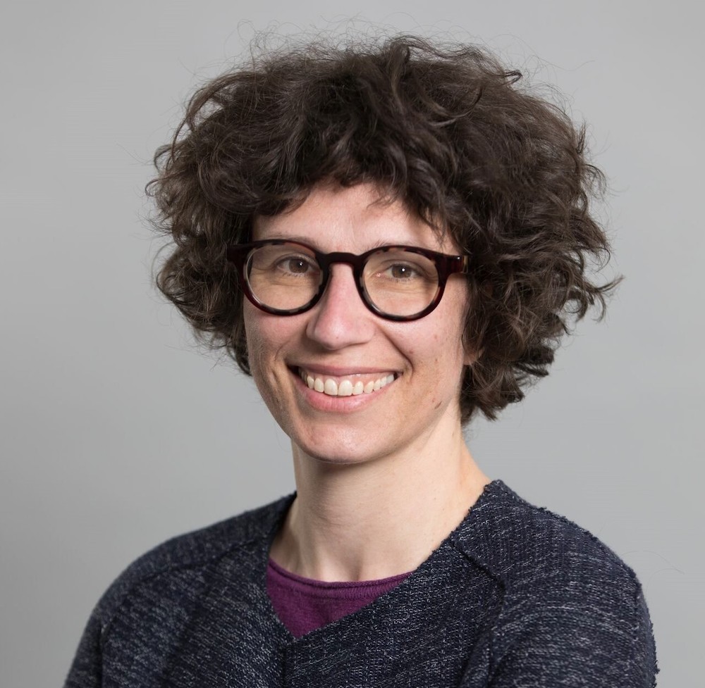 A light-skinned woman with short curly brown hair and thick-rimmed glasses smiles into the camera.