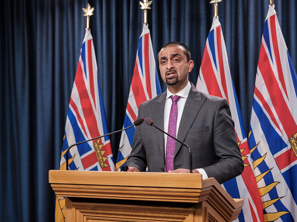 A man with medium skin tone with short dark hair and a goatee stands at a podium. He is wearing a dark suit and lavender tie.