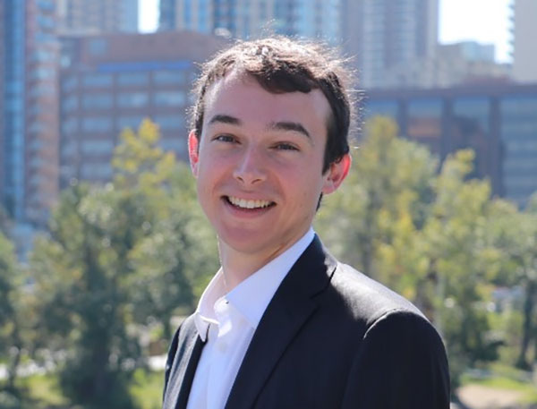 A young light-skinned man with short dark hair stands outdoors and smiles at the camera. He wears a dark suit jacket, white short and no tie.