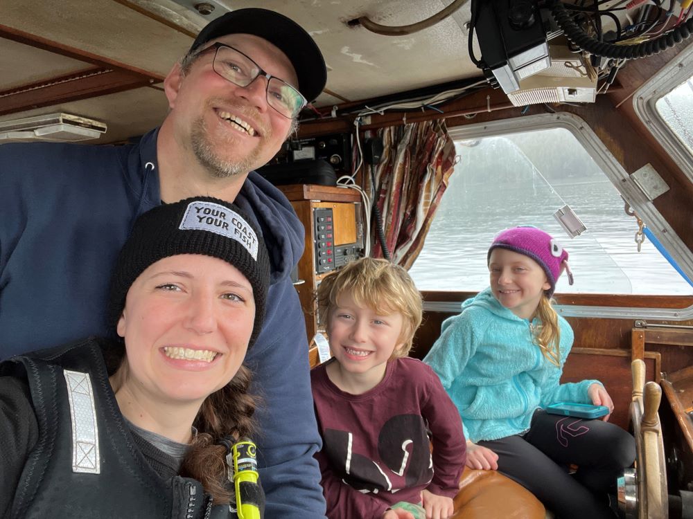 Melissa Collier, centre left, smiles at the camera. She&rsquo;s black wearing a black toque and her brown hair is in a braid. Joel Collier, in a navy hoodie, glasses and black ball cap, stands directly behind her, smiling. Their children are seated to the right of the frame. The family has light skin and hazel or blue eyes. They are seated in the small wood-paneled cabin of a fishing boat.