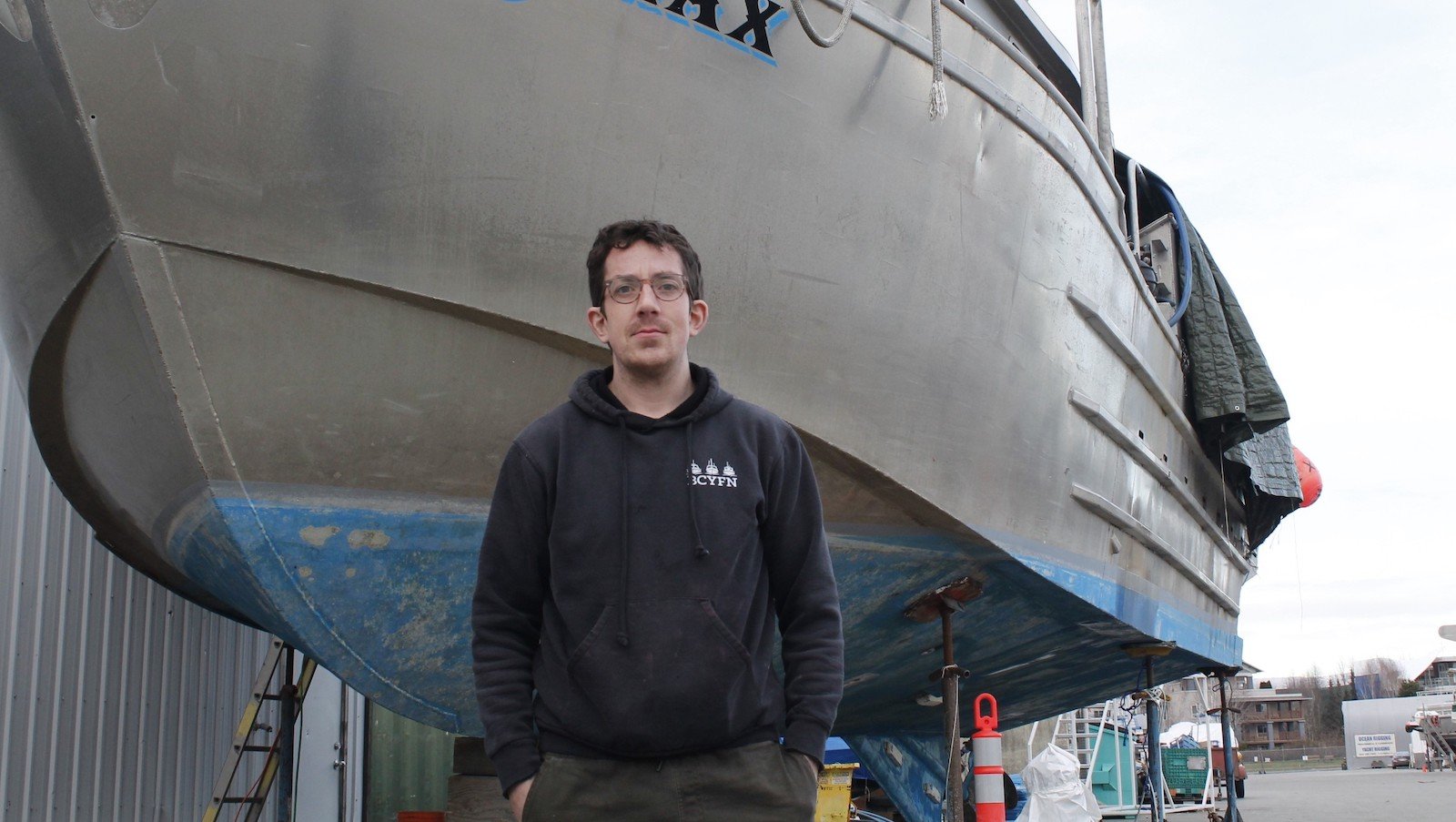 Duncan Cameron has short dark curly hair, light skin and glasses. He is wearing a black hoodie and standing with his hands in his pockets in front of a fishing boat on standers in parking lot. 